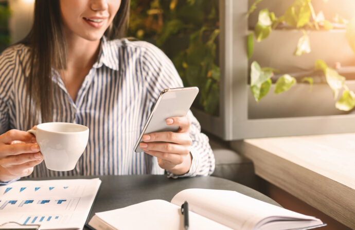 Remote work advantages. Smiling girl chatting on smartphone and drinking coffee, analyzing business plans in cafe, free space (Depositphotos.com photo)