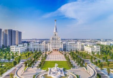 Grand white government building with a tall central spire, flanked by formal gardens and a large plaza with a fountain in the foreground.
