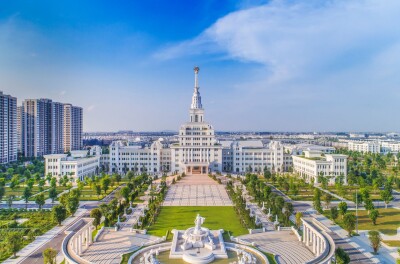 Grand white government building with a tall central spire, flanked by formal gardens and a large plaza with a fountain in the foreground.