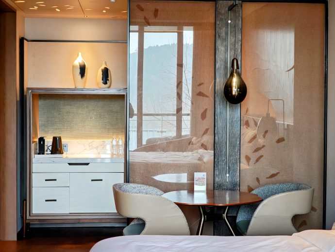 Hotel room seating area with a round wooden table and two curved beige chairs, beside a glass partition with a leaf motif and a dresser with kettle and water bottles.
