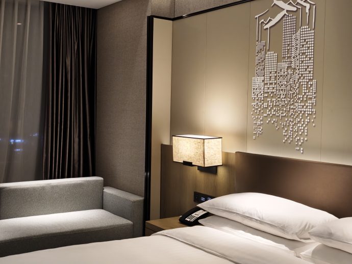 Modern hotel room corner: bed with white linens, bedside lamp, and a gray sofa by a window with drapes.